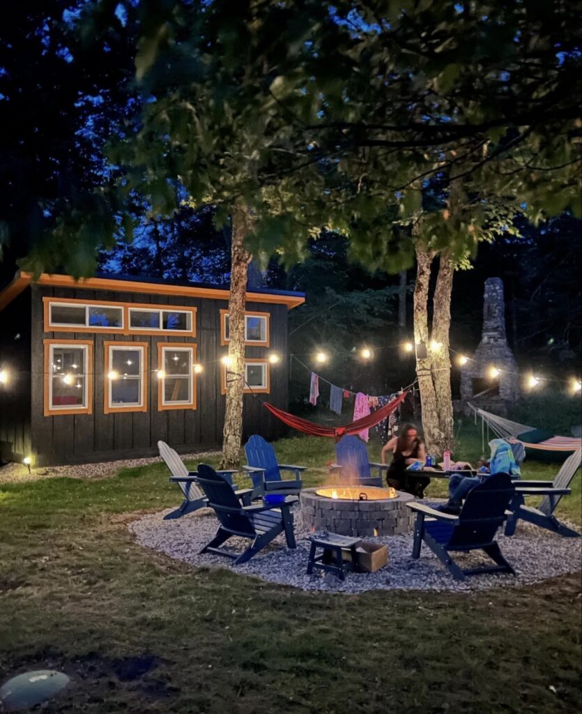 One rustic wooden cabins illuminated at night with warm outdoor string lights, featuring a cozy backyard setup with Adirondack chairs gathered around a lit fire pit.