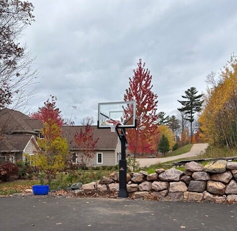 An in-ground 72-inch full-size basketball hoop installed in a residential yard near a rock retaining wall and trees."